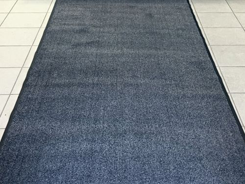 Et teksturert blått Matteutleie-teppe lagt på et hvitt flislagt gulv, som strekker seg over bildet. Teppet virker tykt og slitesterkt, egnet for områder med mye fottrafikk som inngangsmatter. (Merkenavn: Aurskog Bygg & Renhold)