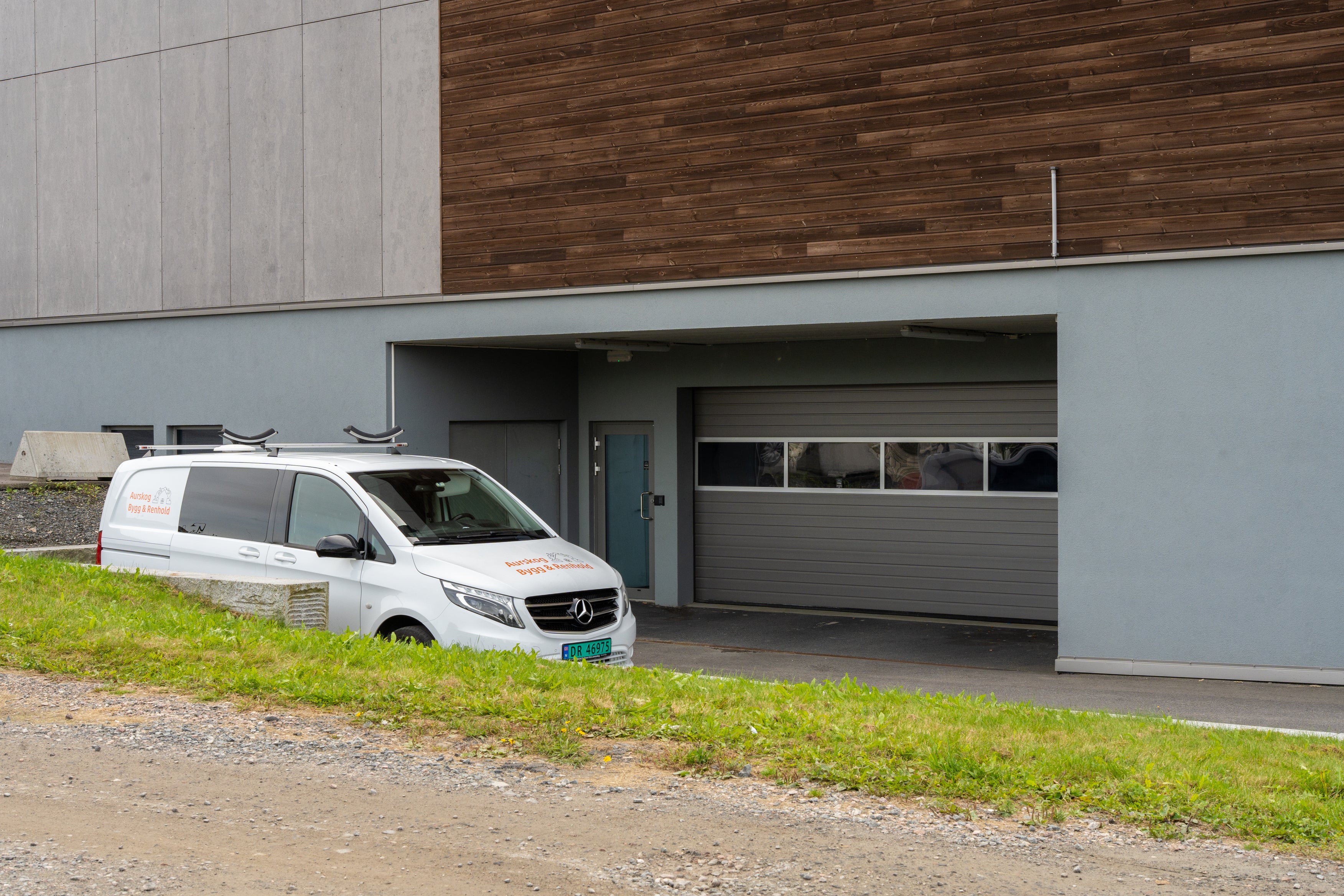 En hvit varebil parkert utenfor et moderne bygg med grå- og trepanelvegger, ved siden av en sammenrullet garasjeport i metall på parkeringsplassene. Det er her Aurskog Bygg & Renholds Feiing finner sted.