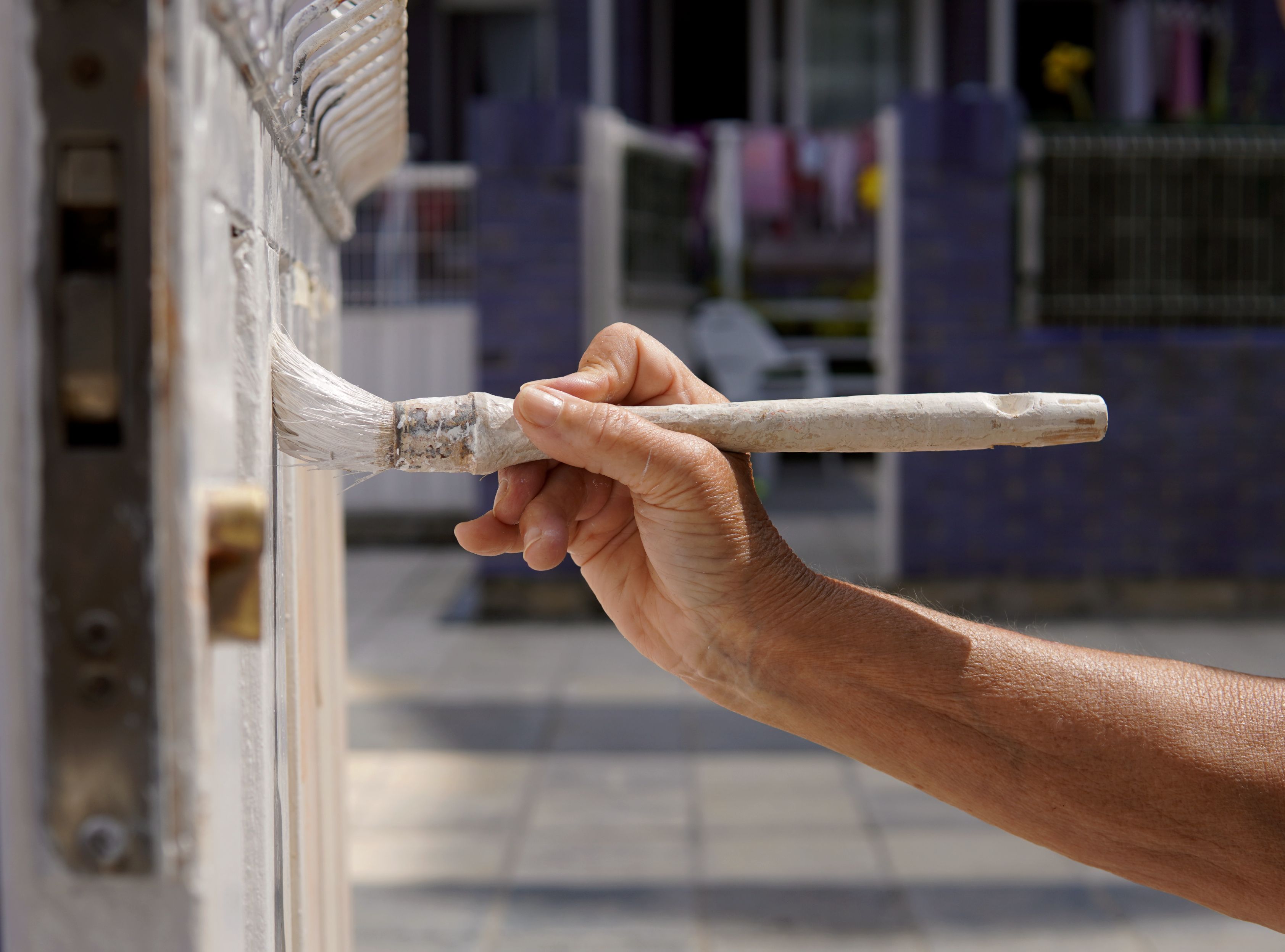 Nærbilde av en persons hånd som holder en pensel, påfører Malerarbeid hvit maling på en metallport i dagslys, med uskarp bakgrunn som viser bedrifter. (Merkenavn: Aurskog Bygg & Renhold)
