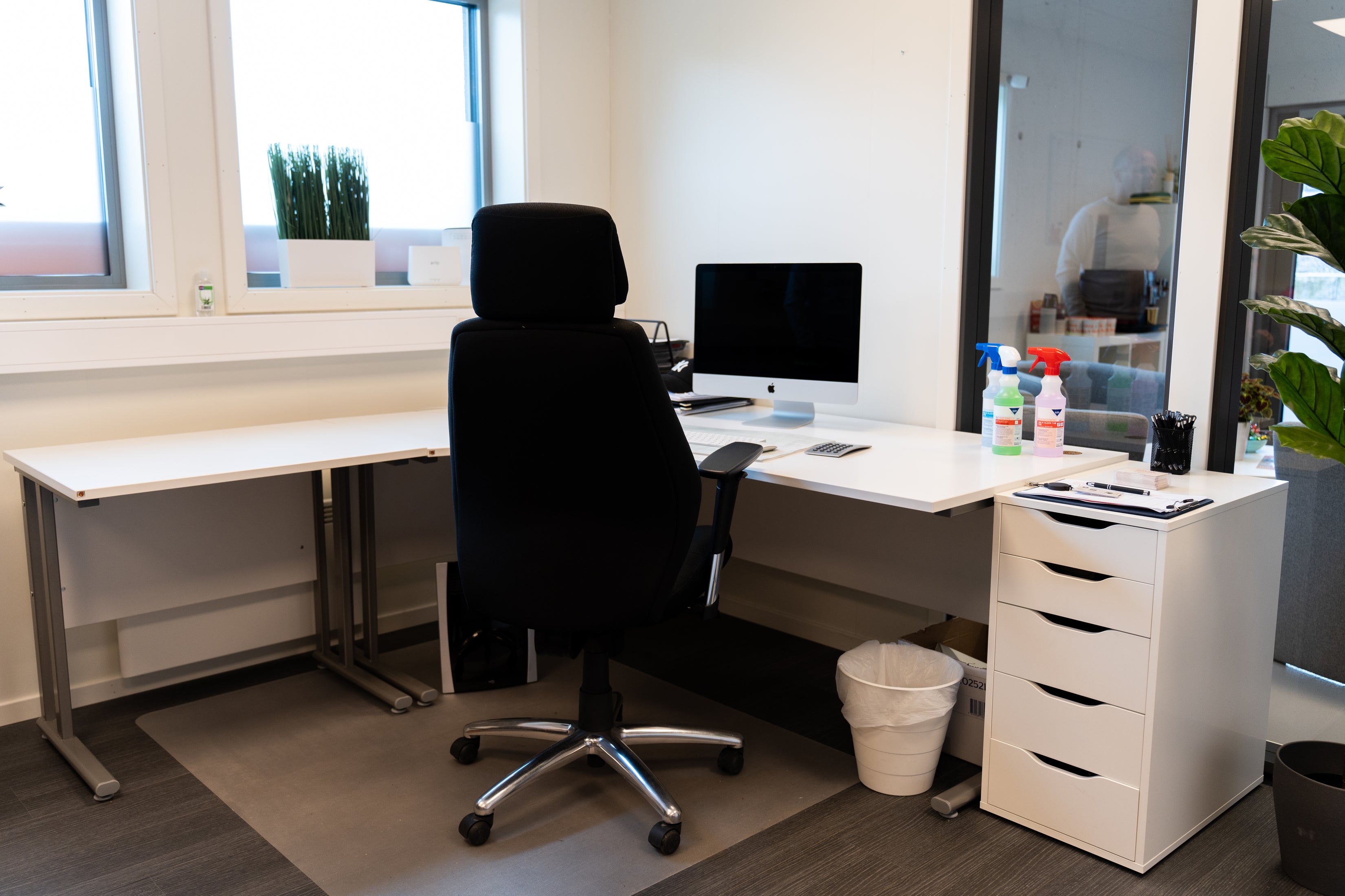 En moderne kontorplass designet for Fast renhold hos bedrifter har en svart kontorstol, et hvitt skrivebord med en dataskjerm, rensemiddel og papirarbeid. En liten søppelbøtte og oppbevaringsenhet er også synlig.